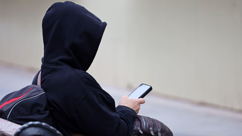 Hombre con sudadera con capucha usando un teléfono inteligente sentado en un banco de una calle de la ciudad