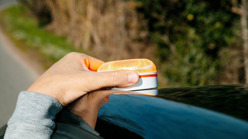 Un hombre coloca una luz de advertencia V16 encendida sobre el techo de su automóvil en una tranquila carretera rural para señalar una avería del vehículo durante el día.