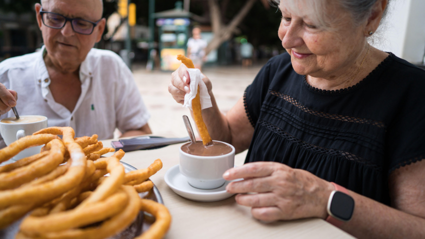 Una pareja de jubilados saboreando churros al aire libre. La mujer moja el churro en chocolate caliente, creando un ambiente acogedor y tradicional. Un plato de churros.