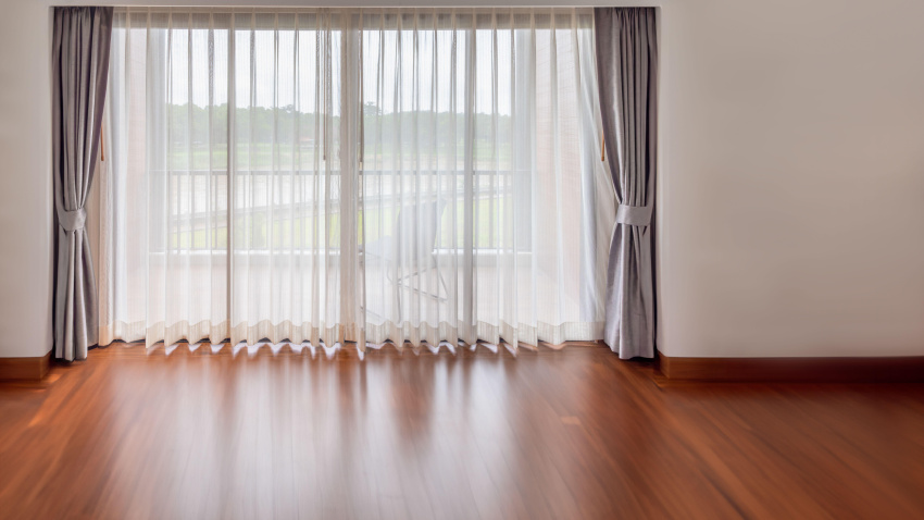 Interior de una sala de estar vacía con una gran ventana luminosa y cortinas blancas en una habitación vacía.