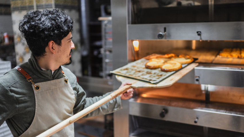 Baker using a wooden peel to remove a tray of golden brown pastries from a professional oven in a bakery