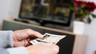 Primer plano de un hombre con algunos billetes de la Lotería de Navidad española en la mano mientras ve la emisión televisada