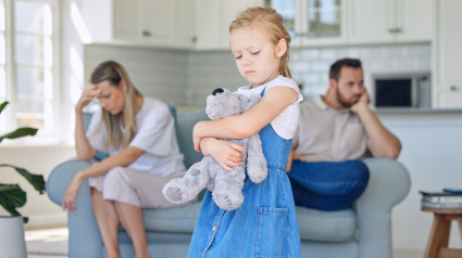 Una niña molesta aprieta su osito de peluche mientras se ve triste y deprimida mientras sus padres discuten de fondo