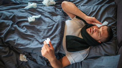 Imagen de un joven acostado en la cama con dolor de cabeza y goteo nasal. Se siente mal y agotado.