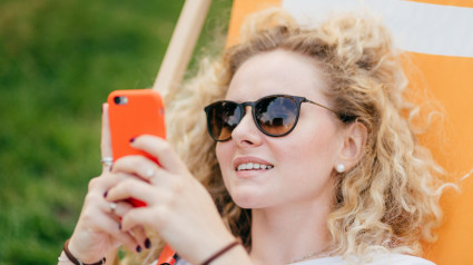 Fotografía al aire libre de una joven de aspecto agradable y rizado con gafas de sol, que sostiene un moderno teléfono inteligente naranja y navega en las redes sociales