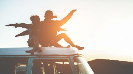 Silueta de pareja feliz sentada en la parte superior de una minivan al atardecer: jóvenes divirtiéndose en las vacaciones de verano viajando alrededor del mundo