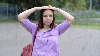 Mujer positiva con cabello corto y camisa morada sosteniendo sus manos sobre su cabeza, protegiéndose de la lluvia