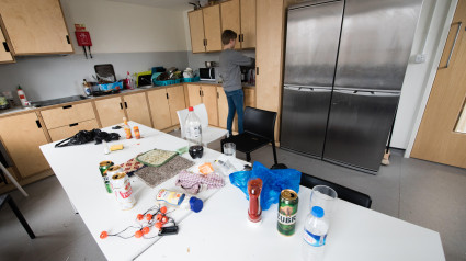 Cocina compartida de estudiantes sucia y desordenada con pilas de platos sucios junto al fregadero y una mesa común llena de basura, latas de cerveza vacías y comida.