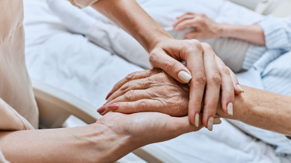 Un pariente sostiene la mano temblorosa de una mujer mayor con enfermedad de Parkinson acostada en una cama de hospital en la sala médica