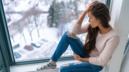 Chica triste deprimida en invierno, sola junto a la ventana de su casa mirando el clima frío, molesta e infeliz