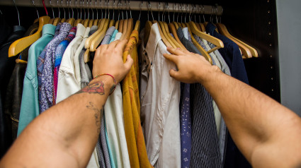 Hombre buscando una camisa colgada en la barra de su armario, vista en primera persona mirando hacia abajo desde sus brazos