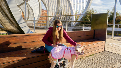 Una mujer joven acaricia a su perro galgo, que lleva un abrigo acolchado rosa, mientras está sentada en un banco en la pasarela de Arganzuela en Madrid