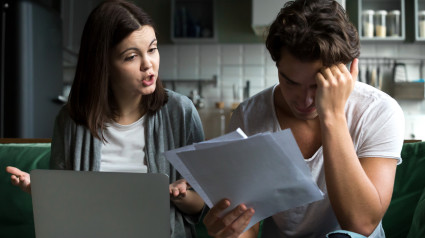 Angry wife scolding blaming upset husband of unpaid bills, bankruptcy or past due debt, unhappy millennial couple having financial problems arguing at home sitting on sofa with laptop and papers