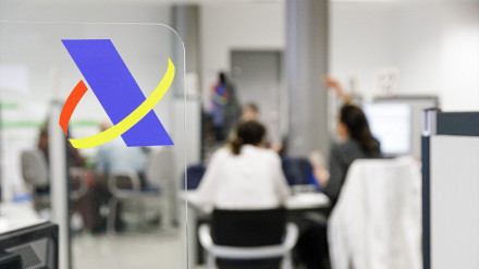 Logotipo de la Agencia Tributaria donde al fondo se ve la oficina con dos personas que están siendo atendidas por un funcionario de Hacienda.