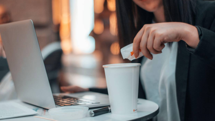 2HCEF4C 2HCEF4C Young manager worker pouring sugar into coffee in a cafeteria in the city square while working with her laptop
