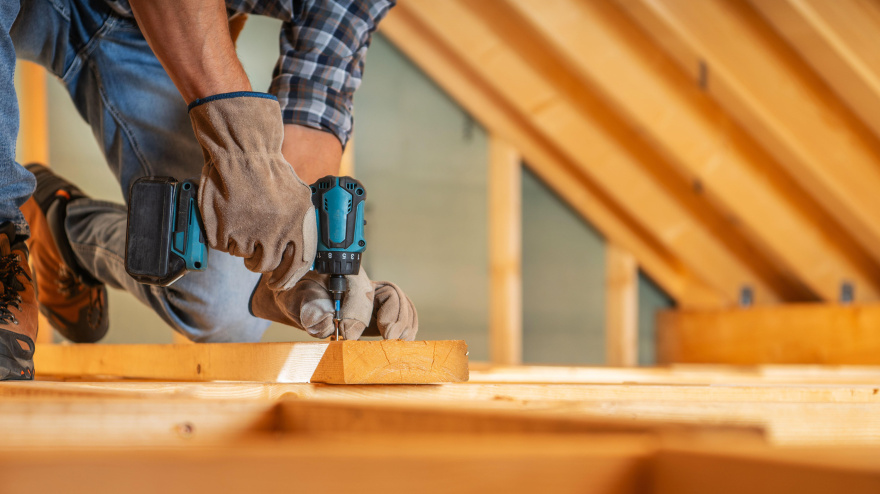 Un carpintero trabaja en un ático, asegurando vigas de madera con un taladro eléctrico mientras usa guantes.