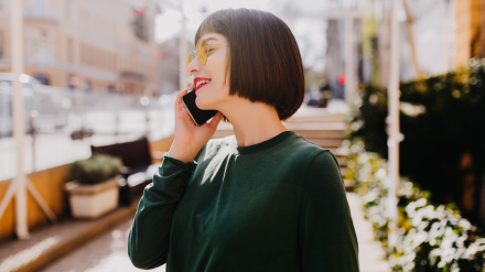 Glamorous woman with short haircut talking on phone. Beautiful brunette girl in green sweater calling someone on street background.