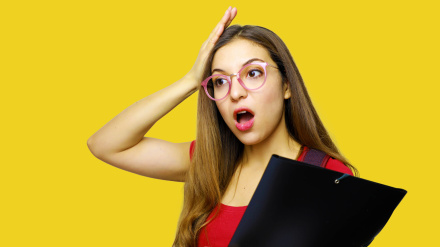 Una joven asustada con la boca abierta se sostiene la cabeza con la mano, lleva gafas y una camiseta roja y lleva una carpeta, se olvida de hacer algo importante.