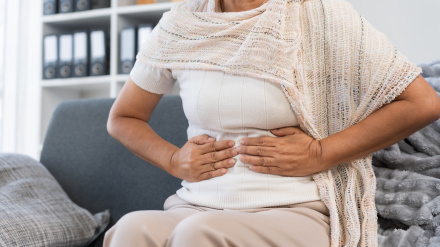 Elderly woman experiencing abdominal pain and discomfort, sitting on a sofa at home, indicating indigestion or health issues