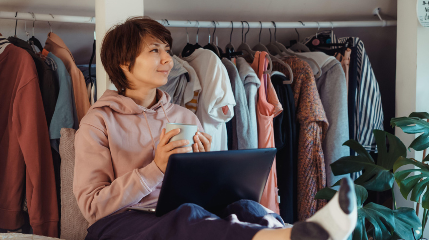 Mujer con una taza de café sentada en la cama, usando el portátil y con el armario abierto