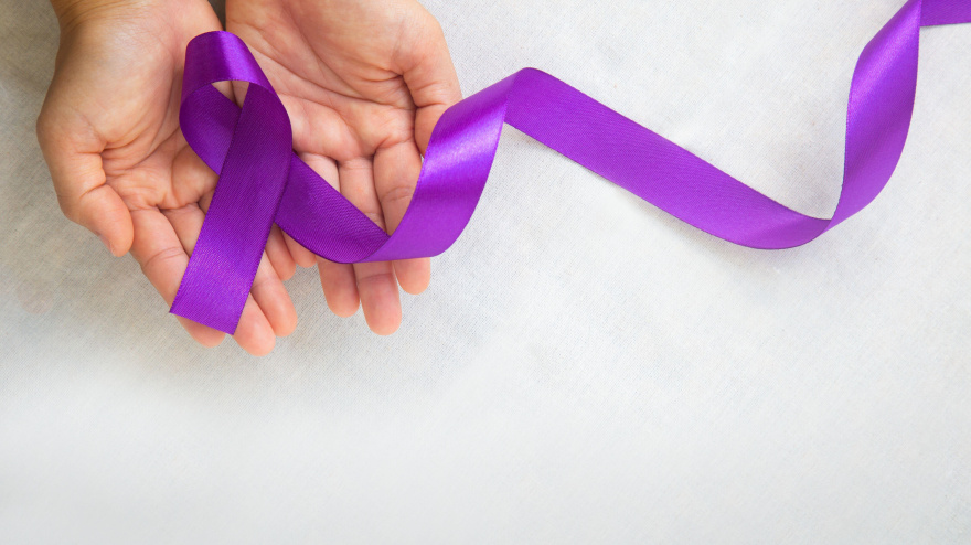 Manos sosteniendo una cinta morada o violeta sobre tela blanca para concienciar sobre el  cáncer