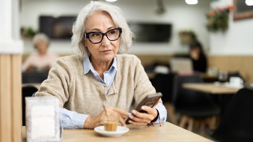 Mujer jubilada bebiendo café recién hecho en la mesa de la cafetería y mirando el teléfono móvil