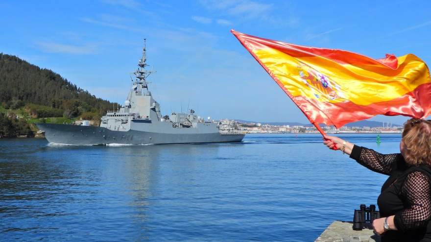 La fragata este domingo saliendo de la ría de Ferrol ante una mujer con una bandera de España