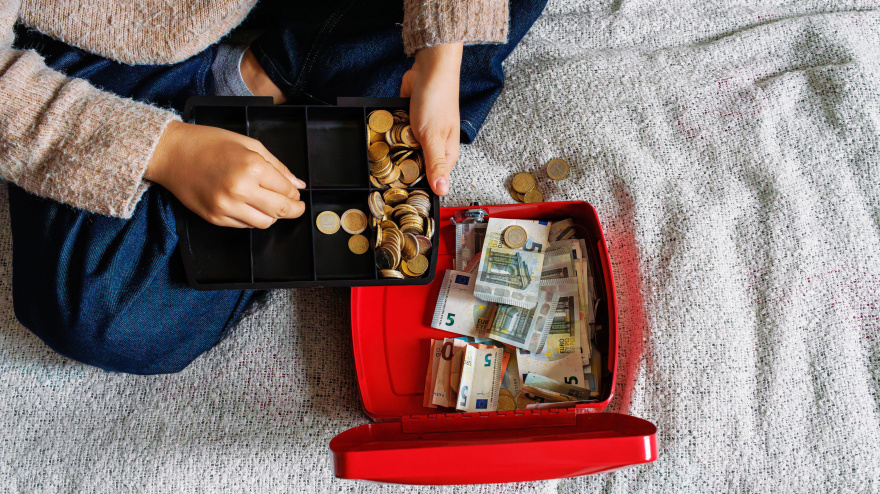 Una joven sentada en una cama cuenta monedas de euro en una caja registradora roja abierta, llena de billetes de euro.