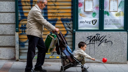 Un jubilado lleva una bolsa de plástico y un cochecito de bebé con un niño que tiene un globo rojo