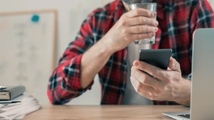 Trabajador autónomo bebiendo agua y usando un teléfono móvil en la oficina de su casa durante un proyecto de trabajo independiente