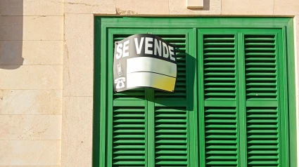 Fachada de una casa con una típica contraventana verde mallorquina, con un cartel de "se vende" escrito en español.