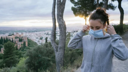 Mujer joven poniéndose mascarilla en la calle para protegerse de virus, enfermedades infecciosas y aire contaminado en la ciudad al atardecer