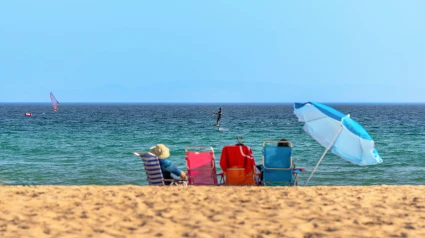 Escena de playa con gente disfrutando del sol sentada en sillas de colores con una sombrilla al lado.