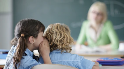 Joven susurra a un compañero en clase delante de la profesora