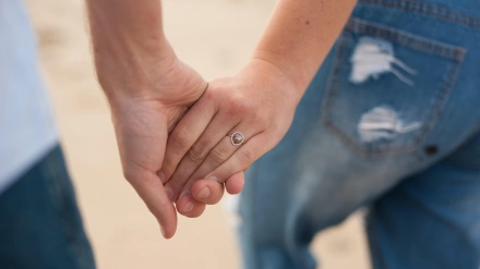 Fotografía horizontal de primer plano de una joven pareja tomada de la mano en la playa con el foco puesto en el anillo durante una sesión de fotos en la playa en un día nublado