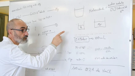 Profesor de unos 50 años con bata de laboratorio explicando química en una pizarra