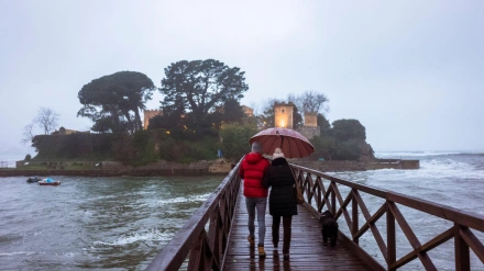 Dos personas con paraguas caminan por la pasarela en dirección a la isla de Santa Cristina, Oleiros, provincia de A Coruña.