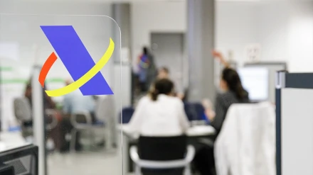 Logotipo de la Agencia Tributaria donde al fondo se ve la oficina con dos personas que están siendo atendidas por un funcionario de Hacienda.