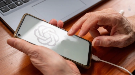 Las manos de un jubilado sosteniendo un teléfono inteligente con el logotipo de ChatGPT en la pantalla sobre un escritorio con una computadora portátil cerca.