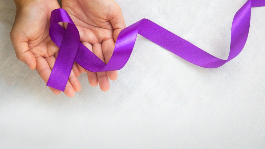 Manos sosteniendo una cinta morada o violeta sobre tela blanca para concienciar sobre el  cáncer