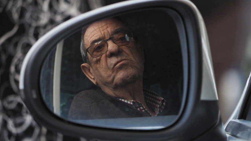 Reflejo de un hombre jubilado, severo y con gafas, visible en el espejo retrovisor exterior de un automóvil.