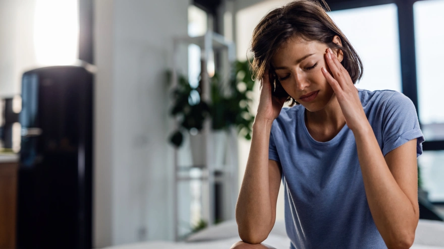 Sad woman suffering from headache and sitting on the bed with eyes closed while holding her head in pain.